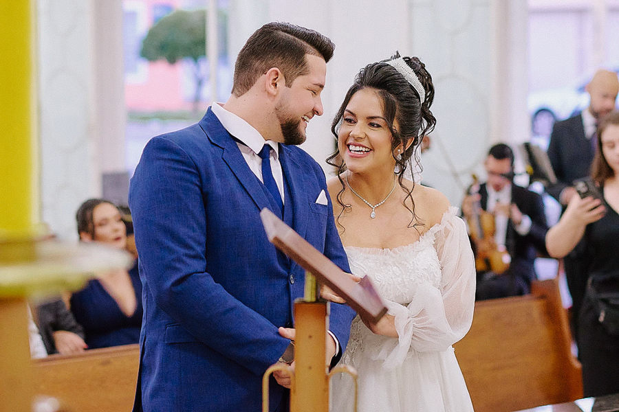 Noivos Paloma e Cassiano trocam olhares apaixonados e sorrisos no altar da Capela Nossa Senhora Aparecida em São José dos Campos. Fotografia documental de casamento capturando a felicidade espontânea do casal durante a celebração religiosa.