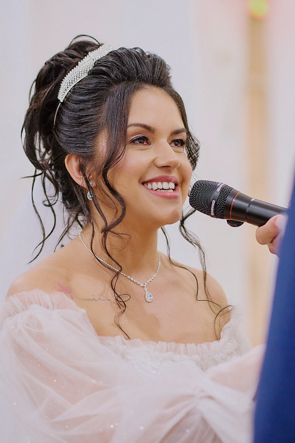 Noiva Paloma sorri emocionada ao falar no microfone durante os votos na Capela Nossa Senhora Aparecida, São José dos Campos. A fotografia destaca sua expressão de felicidade, a tiara brilhante e o colar delicado em um momento de profunda entrega.