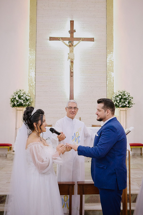 Noivos Paloma e Cassiano no altar da Capela Nossa Senhora Aparecida, em São José dos Campos, durante a troca de alianças conduzida pelo celebrante. Fotografia de casamento com plano aberto destacando o crucifixo e a decoração clássica da igreja.