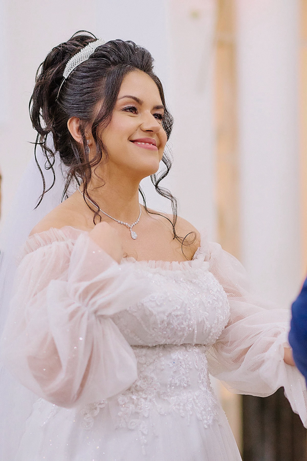Noiva Paloma sorri com olhar de felicidade e realização no altar da Capela Nossa Senhora Aparecida, em São José dos Campos. A fotografia destaca o brilho da tiara e os detalhes delicados das mangas do vestido após a troca de alianças com Cassiano.