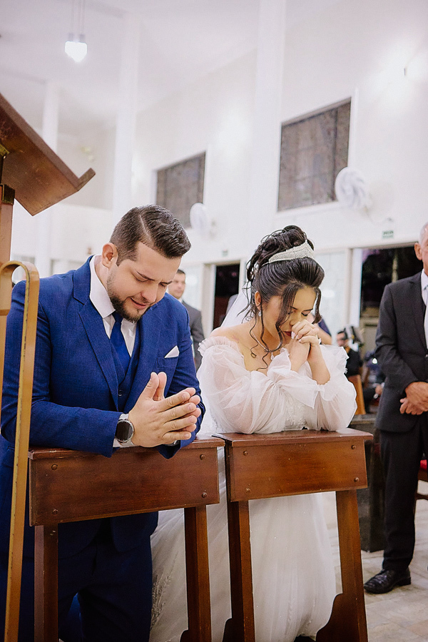 Noivos Cassiano e Paloma ajoelhados em oração no altar da Capela Nossa Senhora Aparecida, em São José dos Campos. Fotografia documental de casamento capturando a espiritualidade, a fé e a união do casal durante a bênção final da cerimônia religiosa.