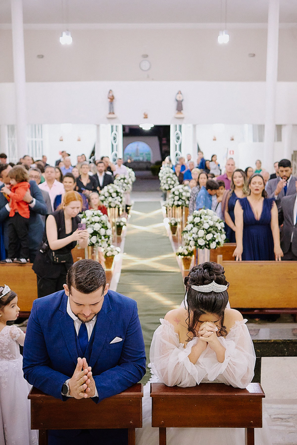 Vista panorâmica dos noivos Cassiano e Paloma ajoelhados em oração no altar da Capela Nossa Senhora Aparecida, em São José dos Campos. A foto captura a nave da igreja repleta de convidados e a atmosfera sagrada do casamento.