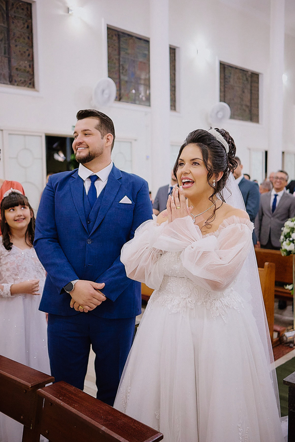 Noivos Paloma e Cassiano sorridentes no altar da Capela Nossa Senhora Aparecida, em São José dos Campos. A noiva expressa alegria com as mãos juntas em celebração, capturando o final emocionante da cerimônia religiosa ao lado de sua família.