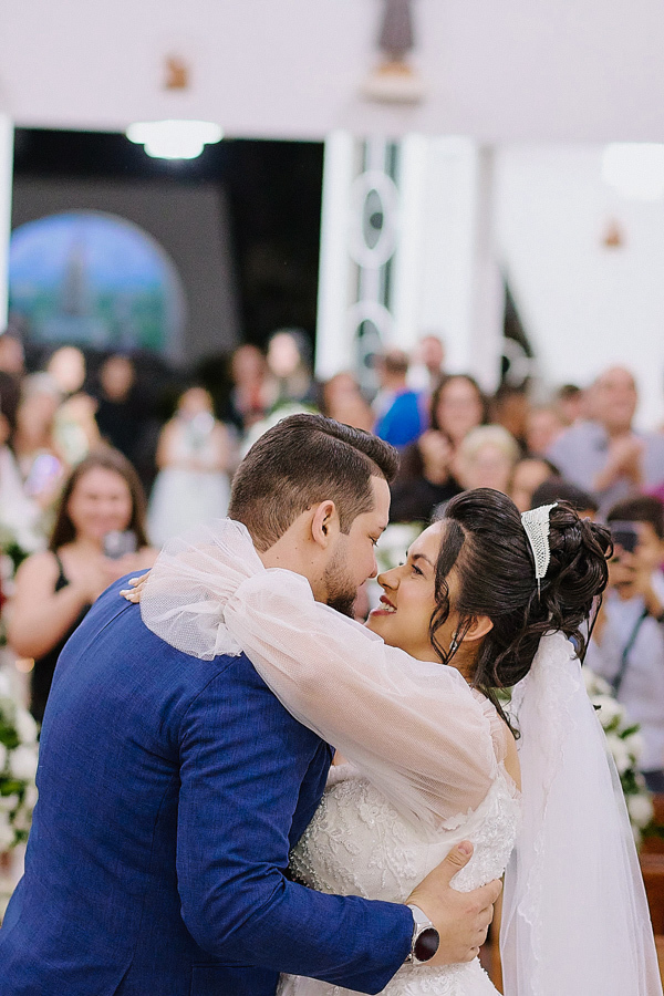 O primeiro beijo de casados de Paloma e Cassiano no altar da Capela Nossa Senhora Aparecida, em São José dos Campos. Fotografia documental capturando o abraço e a felicidade do casal com a igreja repleta de convidados ao fundo.