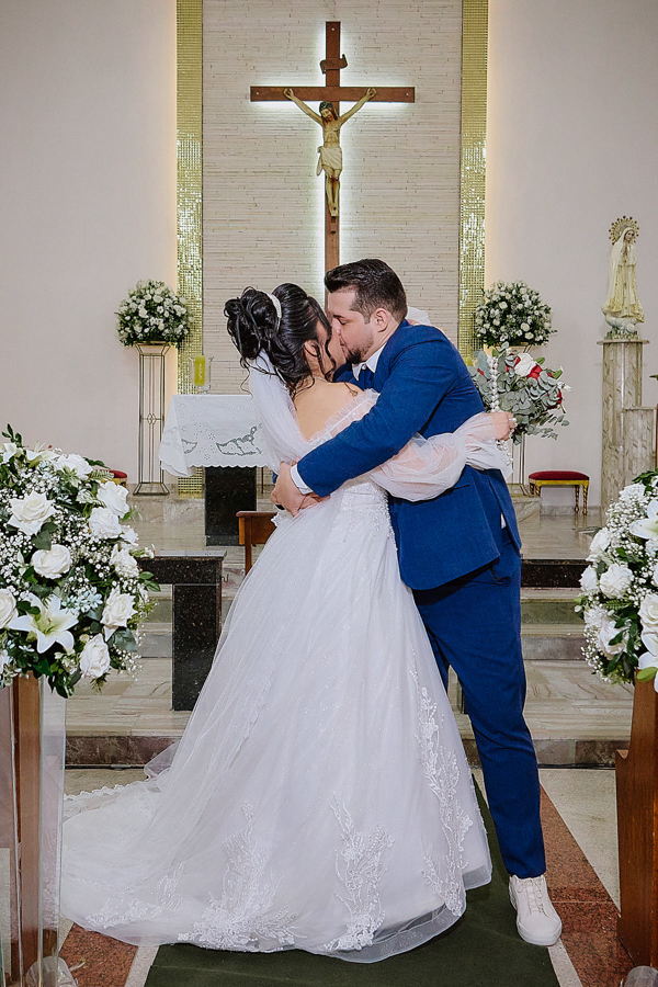 Noivos Paloma e Cassiano trocam o primeiro beijo como casados no altar da Capela Nossa Senhora Aparecida, em São José dos Campos. Fotografia de casamento em plano aberto destacando a cruz, o abraço do casal e a celebração da união familiar.