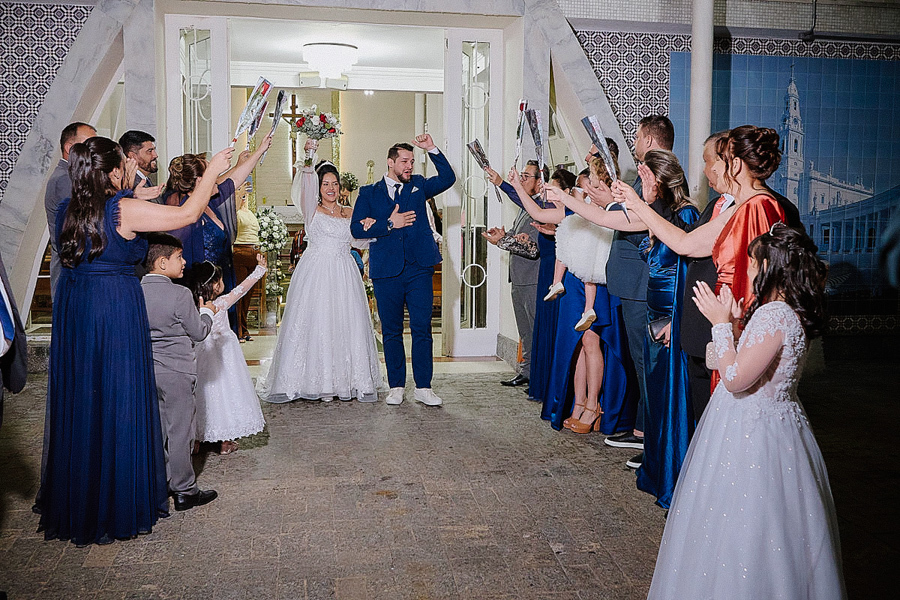 Noivos Paloma e Cassiano saem radiantes da Capela Nossa Senhora Aparecida, em São José dos Campos. Convidados formam um corredor com flores para celebrar o casal e suas filhas em um momento de grande alegria na saída da cerimônia de casamento.