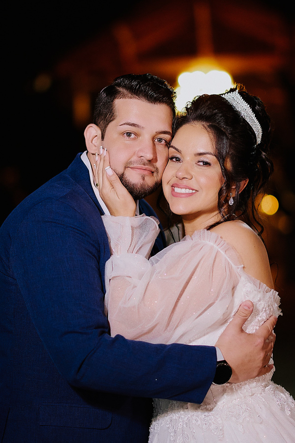 Noiva Paloma acaricia o rosto de Cassiano em um retrato romântico noturno em São José dos Campos. A foto destaca o sorriso dos recém-casados, a tiara brilhante da noiva e a felicidade do casal após o sim na Capela Nossa Senhora Aparecida.