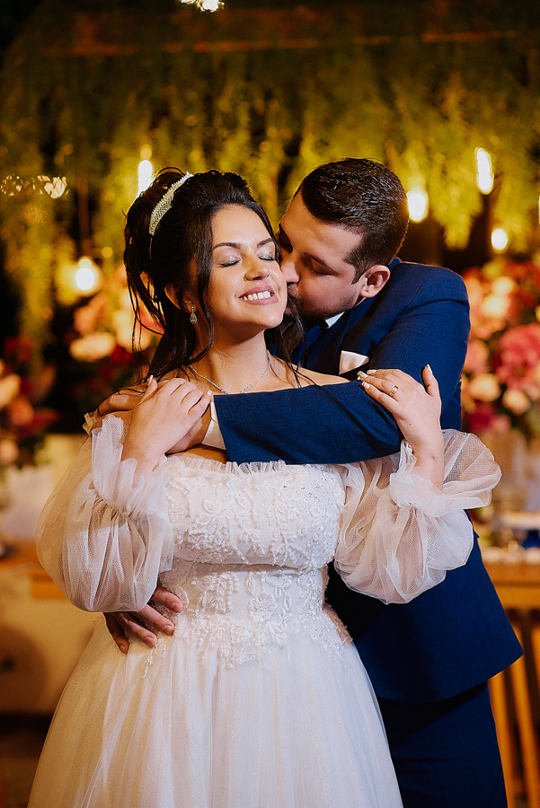 Noivo Cassiano abraça e beija o rosto de Paloma em São José dos Campos. A noiva sorri de olhos fechados, celebrando a união em um cenário romântico com luzes decorativas ao fundo, capturando a felicidade plena do casal após o casamento na Capela.