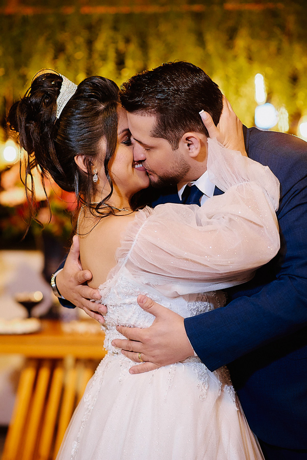 Close romântico dos noivos Paloma e Cassiano trocando um beijo em São José dos Campos. A fotografia destaca o detalhe da manga bufante do vestido de noiva e a felicidade do casal em meio à decoração iluminada da recepção noturna.