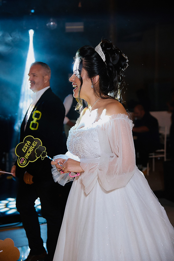 Noiva Paloma sorri animada na pista de dança durante sua festa de casamento em São José dos Campos. Ela segura uma placa divertida 