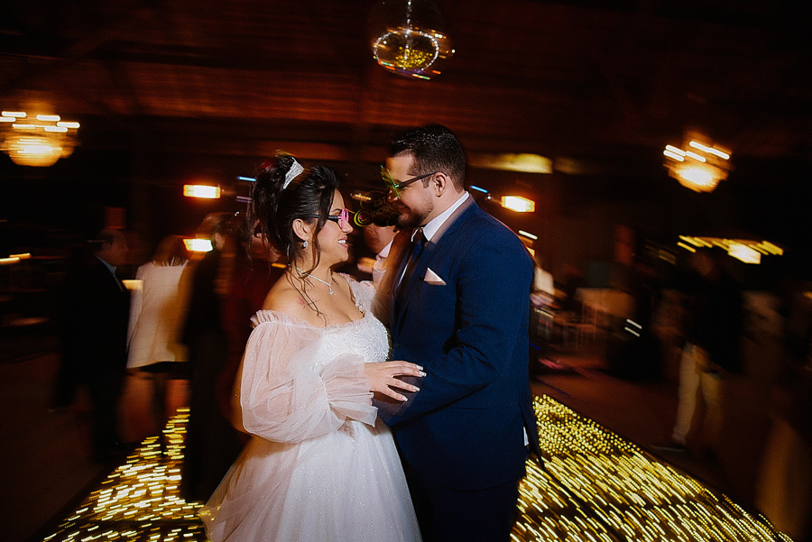 Noivos Paloma e Cassiano dançam felizes na pista de LED durante a festa em São José dos Campos. A fotografia utiliza técnica de longa exposição para criar um efeito de movimento, destacando a alegria e os óculos divertidos do casal na recepção.