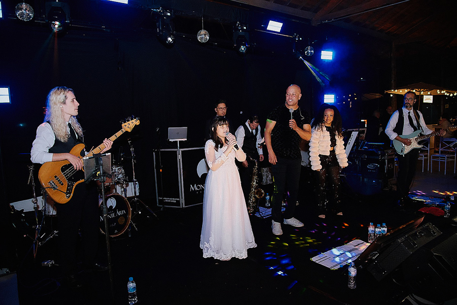 Daminha de casamento canta com a banda ao vivo no palco da Chácara Parque dos Príncipes, em São José dos Campos. Fotografia documental capturando o talento e a alegria da filha de Paloma e Cassiano durante a recepção noturna.