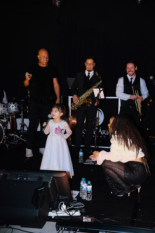 Daminha de casamento canta com microfone no palco durante a festa em São José dos Campos. A fotografia registra a espontaneidade da filha de Paloma e Cassiano, acompanhada pela banda na Chácara Parque dos Príncipes, em um momento fofo da recepção.