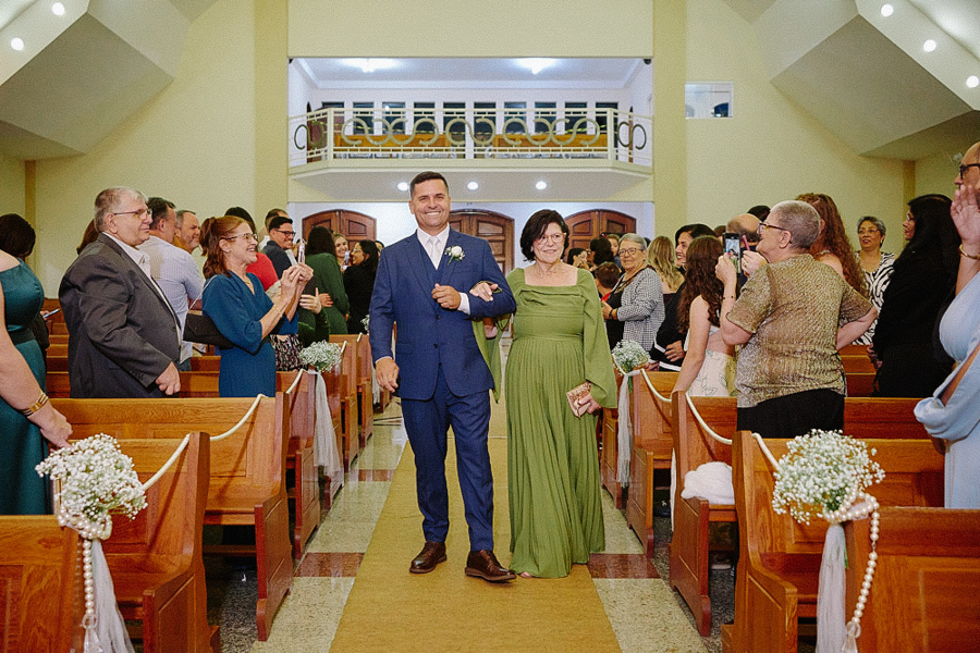 Noivo Fernando entrando sorridente na Igreja Santa Maria em Jacareí de braços dados com a mãe. Fotografia de casamento religioso com convidados ao fundo registrando o momento emocionante da cerimônia.