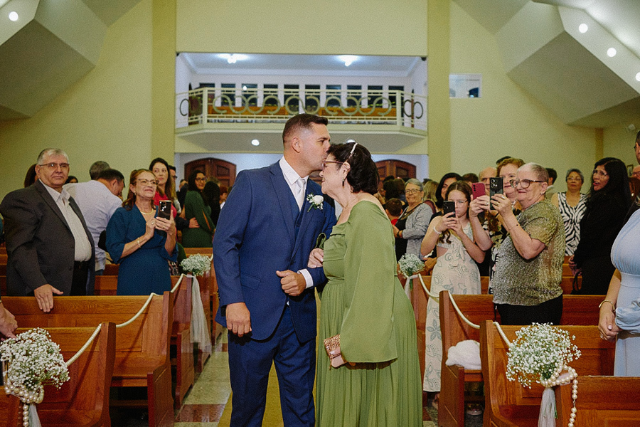 Noivo Fernando beija carinhosamente a testa de sua mãe no corredor da Igreja Santa Maria. Momento de emoção e afeto familiar registrado durante cerimônia de casamento em Jacareí, SP.