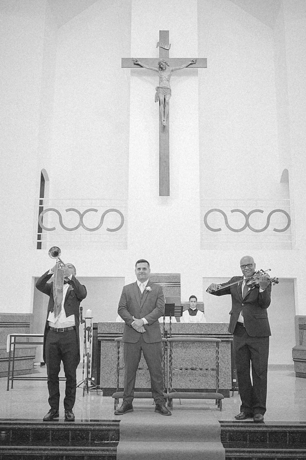 Foto em preto e branco do noivo Fernando aguardando no altar da Igreja Santa Maria em Jacareí, ladeado por músicos tocando trompete e violino. Registro artístico e solene do início da cerimônia de casamento religioso.
