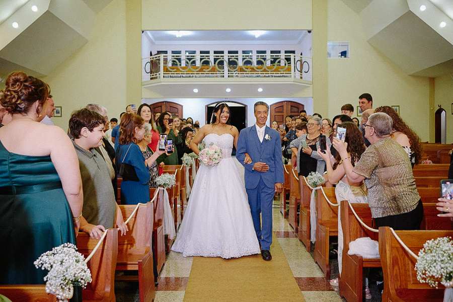 Entrada radiante da noiva Marcela na Igreja Santa Maria em Jacareí. Ela caminha sorrindo pelo corredor central de braços dados com o pai, cercada por convidados registrando o momento. Fotografia de casamento emocionante e espontânea.