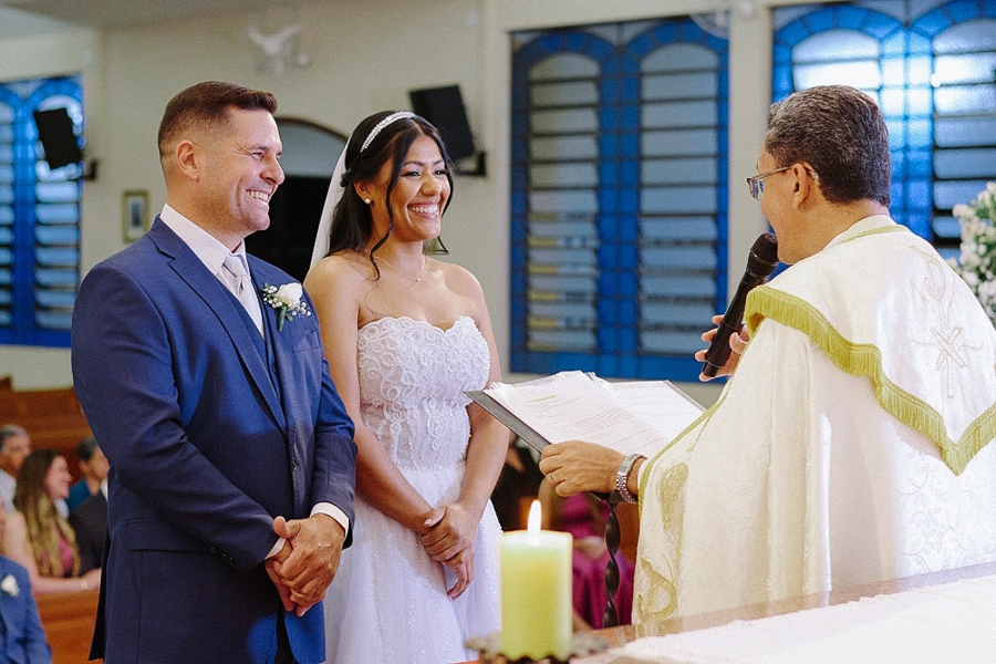 Noivos Marcela e Fernando sorrindo radiantes no altar da Igreja Santa Maria em Jacareí. O casal se diverte com as palavras do padre durante a cerimônia de casamento religioso. Momento de leveza e alegria espontânea.