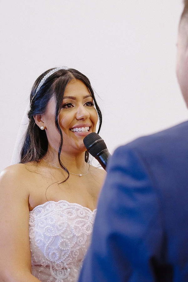 Close da noiva Marcela sorrindo emocionada enquanto diz seus votos no microfone para o noivo Fernando. Detalhe da expressão de felicidade durante a cerimônia de casamento na Igreja Santa Maria em Jacareí.