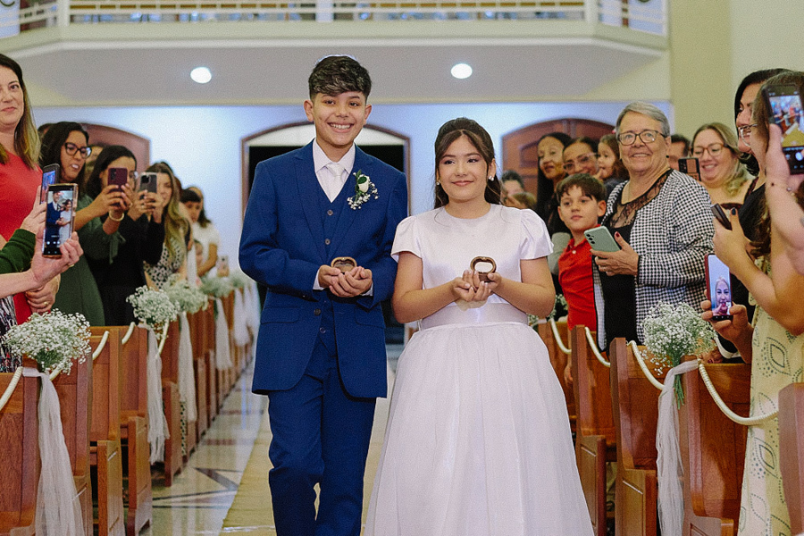 Filhos do casal Marcela e Fernando entram sorrindo na nave da Igreja Santa Maria em Jacareí carregando as alianças. O menino veste terno azul e a menina vestido claro. Momento emocionante do casamento com convidados registrando a cena.
