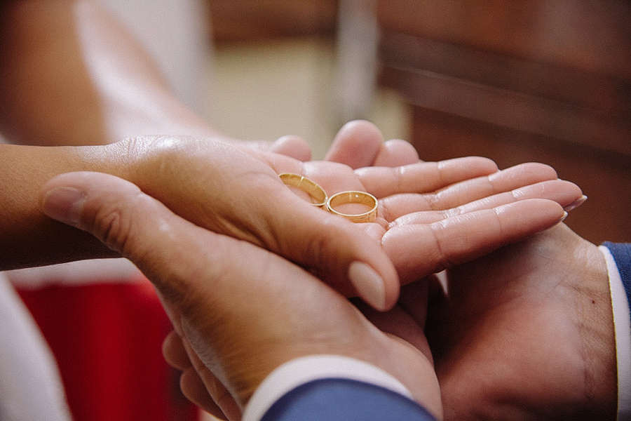 Close-up emocionante das mãos dos noivos Marcela e Fernando segurando as alianças de ouro antes da troca. Detalhe simbólico fotografado durante o casamento religioso na Igreja Santa Maria em Jacareí.