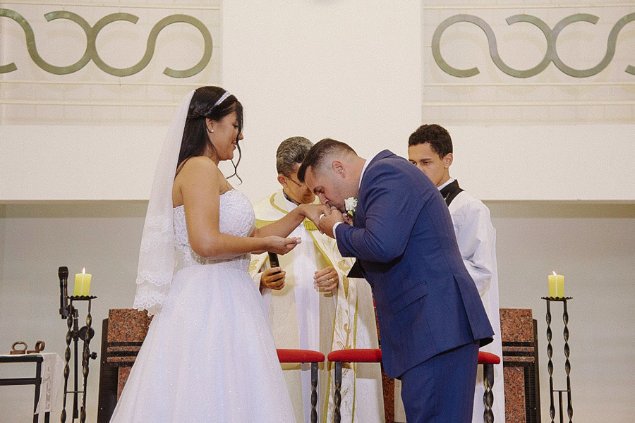 Noivo Fernando beija a mão da noiva Marcela após a troca de alianças no altar da Igreja Santa Maria em Jacareí. Gesto de carinho e respeito durante a cerimônia de casamento religioso.