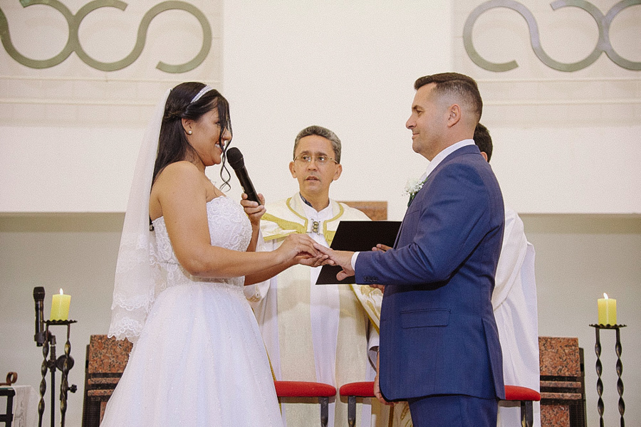 Noiva Marcela coloca a aliança no dedo do noivo Fernando enquanto diz os votos no microfone. Momento solene da troca de alianças no altar da Igreja Santa Maria em Jacareí sob o olhar do padre.