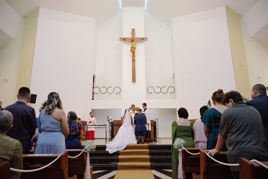 Noivos Marcela e Fernando ajoelhados no altar da Igreja Santa Maria em Jacareí recebendo a bênção. Vista da nave da igreja com convidados em pé e uma grande cruz ao fundo.