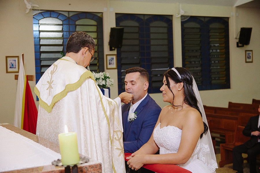 Noivo Fernando recebendo a comunhão das mãos do padre no altar da Igreja Santa Maria em Jacareí. A noiva Marcela aguarda ajoelhada ao lado. Momento solene da eucaristia registrado em fotografia de casamento religioso.