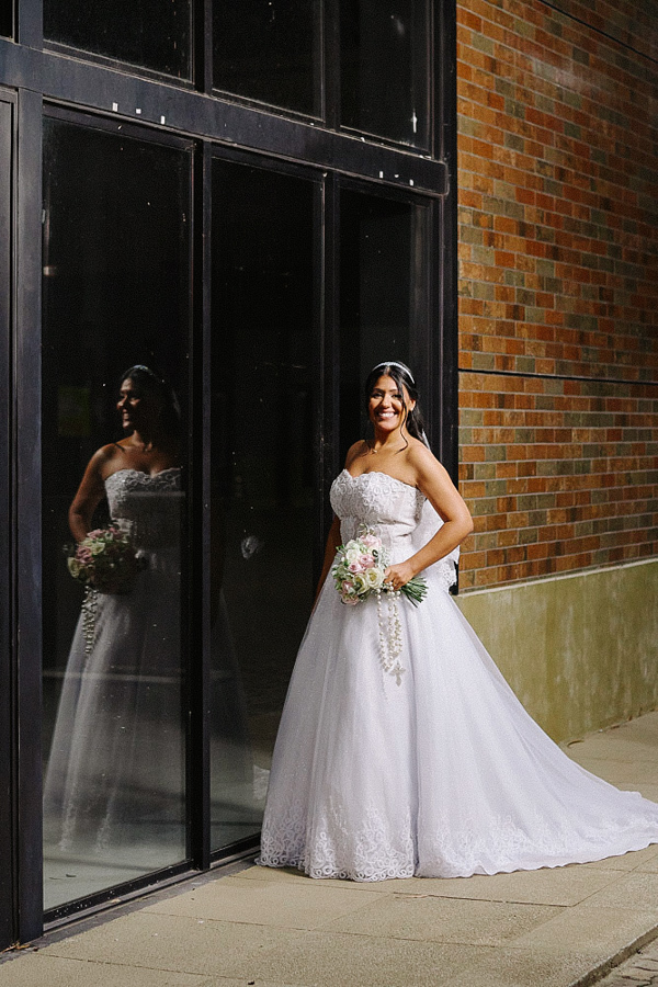 Retrato da noiva Marcela sorrindo radiante, segurando buquê e terço, encostada em uma parede de tijolos com reflexo no vidro. Ensaio de casamento noturno em Jacareí destacando a elegância do vestido de noiva.