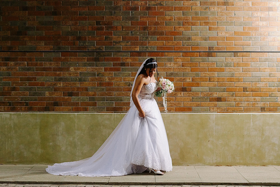 Noiva Marcela caminha elegantemente segurando a saia do vestido e buquê diante de uma parede de tijolos. Ensaio de casamento externo e urbano em Jacareí, destacando o movimento e o visual da noiva.