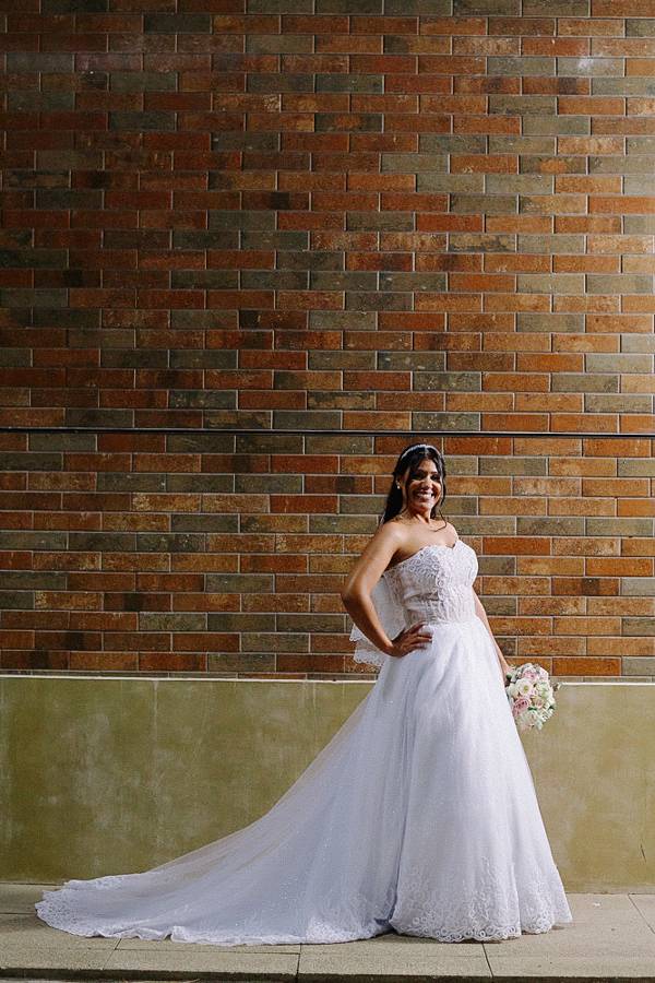 Noiva Marcela posa sorridente com a mão na cintura diante de uma parede de tijolos rústica. Ela usa um vestido tomara que caia com cauda longa e segura o buquê. Ensaio fotográfico de casamento em Jacareí destacando a elegância da noiva.