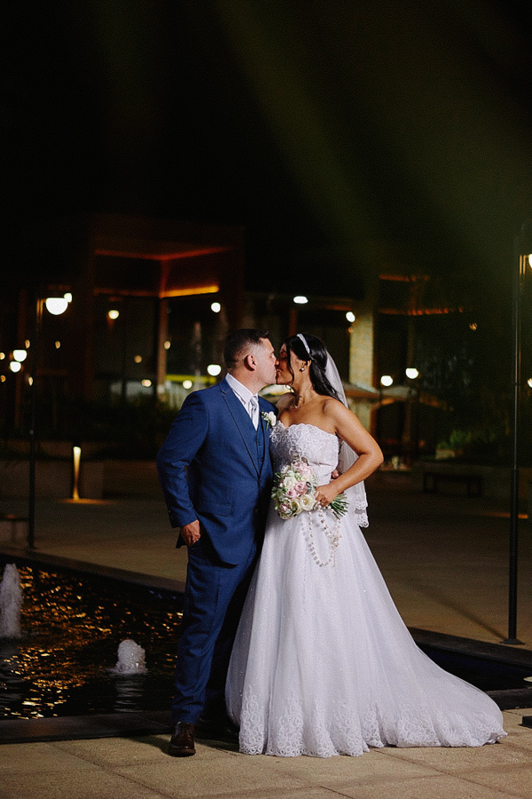 Beijo apaixonado dos recém-casados Marcela e Fernando em ensaio noturno ao ar livre em Jacareí. O casal posa diante de uma fonte iluminada, criando uma cena romântica. Fotografia de casamento externa e elegante.