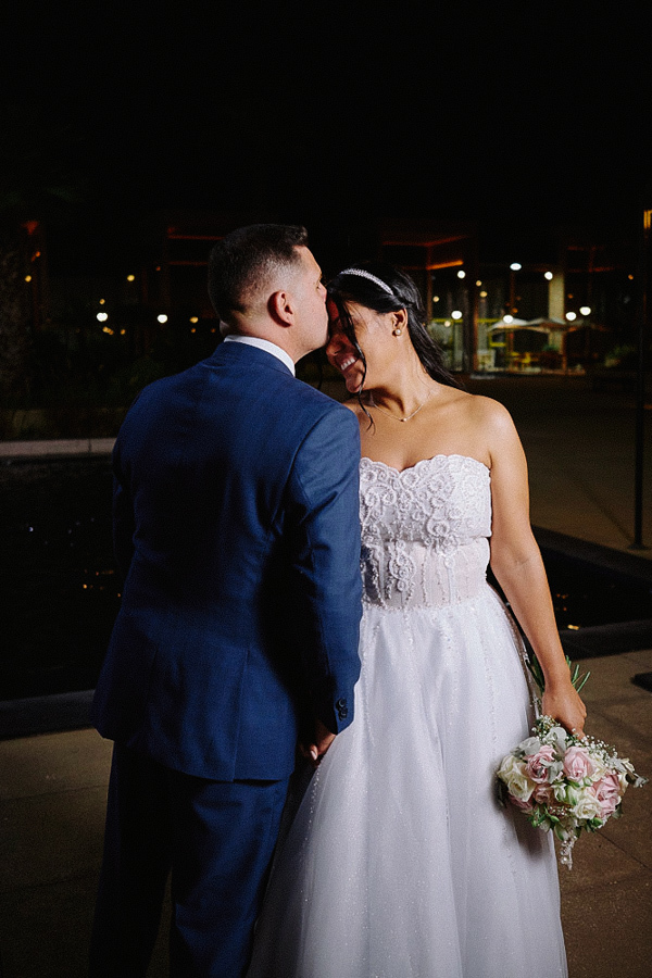 Noivo Fernando beija carinhosamente a testa da noiva Marcela durante ensaio fotográfico noturno em Jacareí. A noiva sorri de olhos fechados segurando o buquê. Fotografia de casamento romântica ao ar livre.