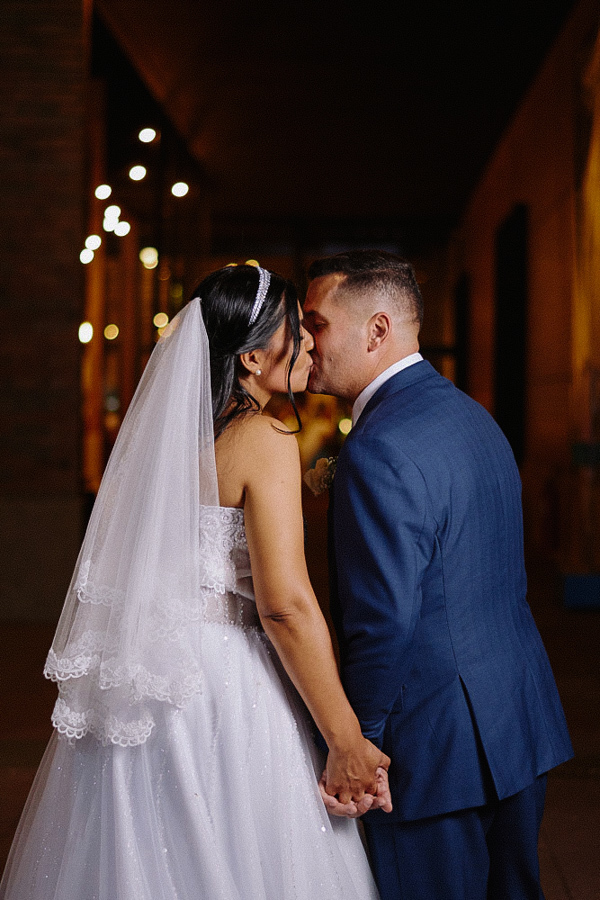 Noivos Marcela e Fernando se beijando apaixonadamente de mãos dadas durante ensaio noturno. O casal está em um corredor com luzes desfocadas ao fundo, destacando o romantismo da fotografia de casamento em Jacareí.