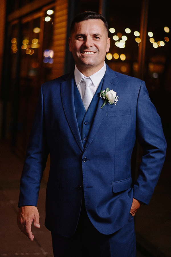 Retrato elegante do noivo Fernando sorrindo para a câmera durante ensaio de casamento noturno em Jacareí. Ele veste terno azul clássico e lapela com rosa branca, com luzes desfocadas ao fundo. Fotografia de noivo espontânea.