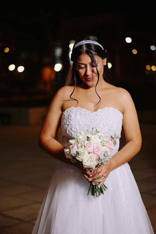 Noiva Marcela contempla seu buquê de rosas em retrato noturno elegante. Ela usa vestido tomara que caia e tiara, com luzes desfocadas da cidade ao fundo criando um bokeh suave. Fotografia de casamento em Jacareí.