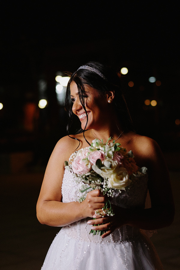 Noiva Marcela sorri radiante segurando seu buquê de rosas com as duas mãos em ensaio noturno. A iluminação destaca sua expressão de felicidade com luzes desfocadas ao fundo. Fotografia de casamento em Jacareí capturando momento espontâneo.
