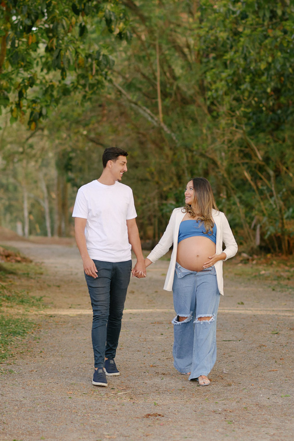 Ensaio fotográfico gestante em São José dos Campos. Lorrana e Felipe caminham sorridentes e de mãos dadas no Parque da Cidade. Ela exibe a barriga à espera de Pedro em um cenário natural com árvores ao fundo.