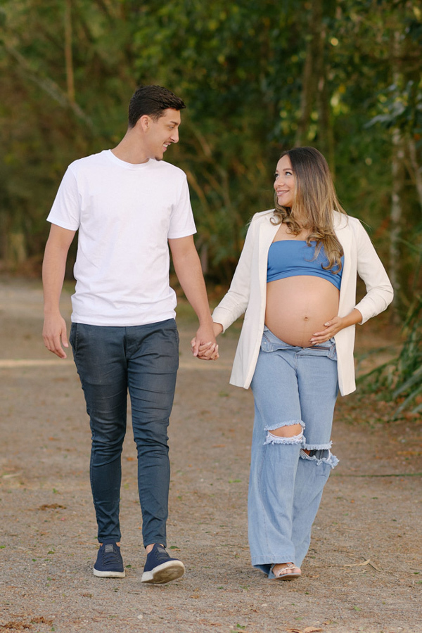 Ensaio fotográfico gestante em São José dos Campos. Lorrana e Felipe caminham sorridentes e de mãos dadas no Parque da Cidade. Ela exibe a barriga à espera de Pedro em um cenário natural com árvores ao fundo, trazendo leveza e amor.