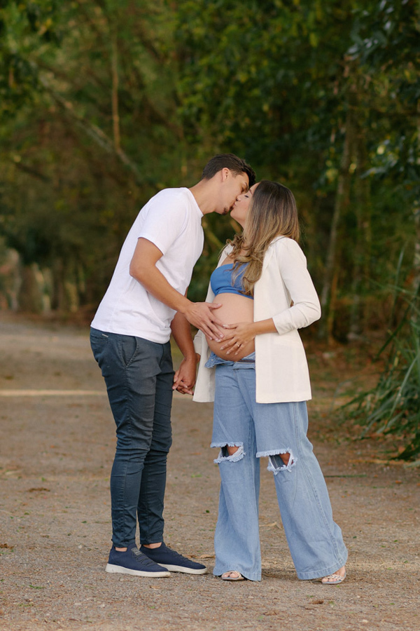 Fotografia de gestante em São José dos Campos. Felipe beija Lorrana enquanto acaricia sua barriga à espera de Pedro. O casal está no Parque da Cidade, cercado pela natureza, em um momento de carinho, conexão e leveza durante o ensaio ao ar livre.