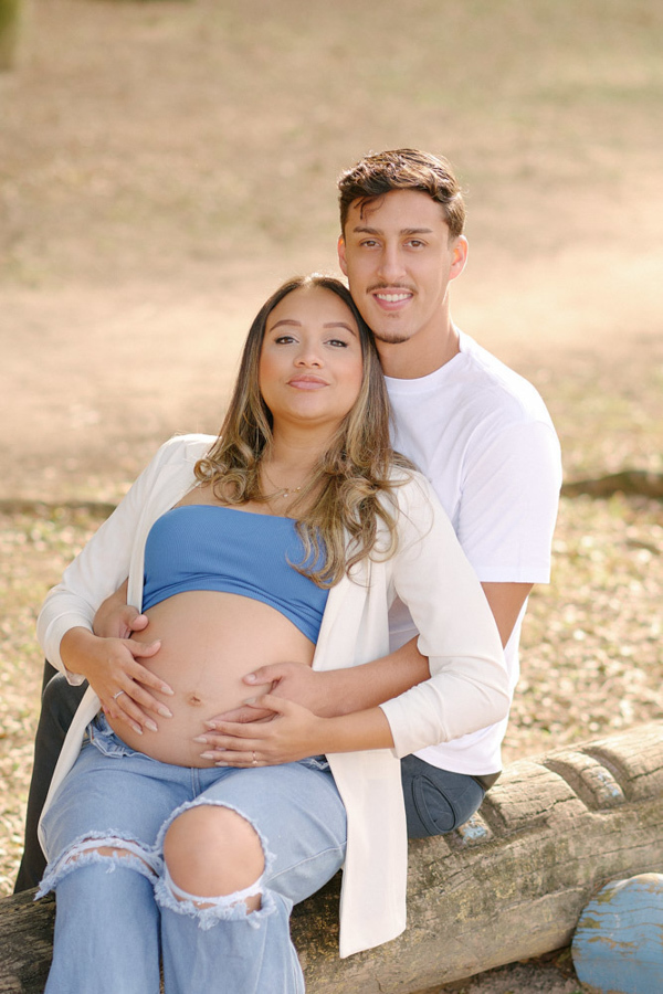 Fotografia de gestante em São José dos Campos. Lorrana e Felipe sentados em um tronco no Parque da Cidade. Ele a abraça por trás enquanto ambos seguram a barriga à espera de Pedro. Um registro íntimo e atemporal de maternidade ao ar livre.