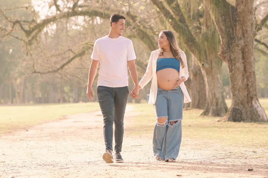 Ensaio gestante em São José dos Campos. Lorrana e Felipe caminham de mãos dadas sob a luz suave do entardecer no Parque da Cidade. A composição destaca a barriga à espera de Pedro e a harmonia do casal em meio às árvores, com um tom quente e acolhedor.