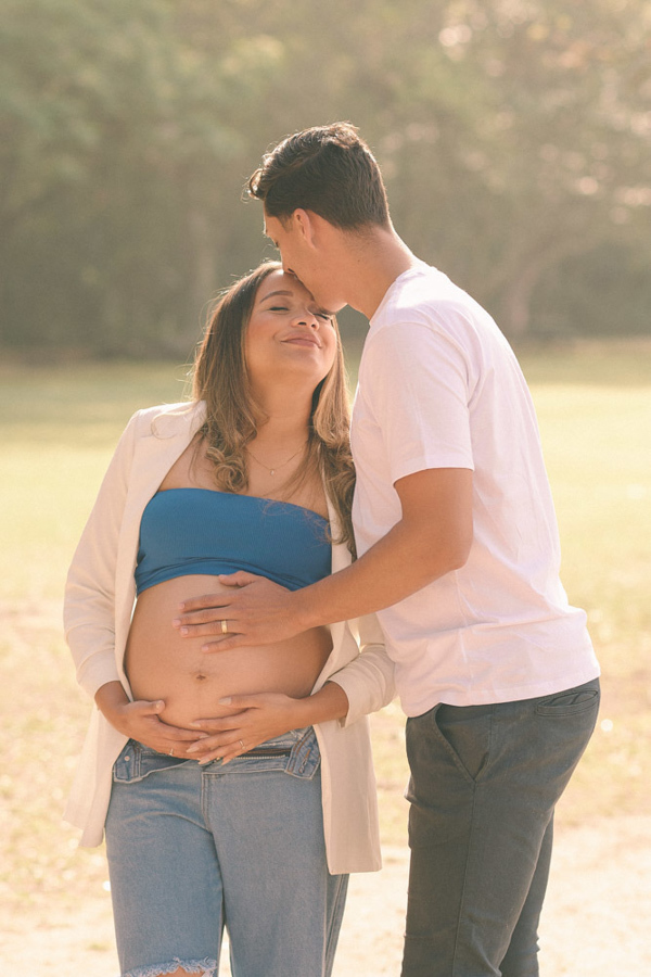 Fotografia de gestante em São José dos Campos. Felipe beija a testa de Lorrana enquanto ambos abraçam a barriga à espera de Pedro. O casal está banhado por uma luz dourada e suave no Parque da Cidade, transmitindo paz e carinho no ensaio ao ar livre.