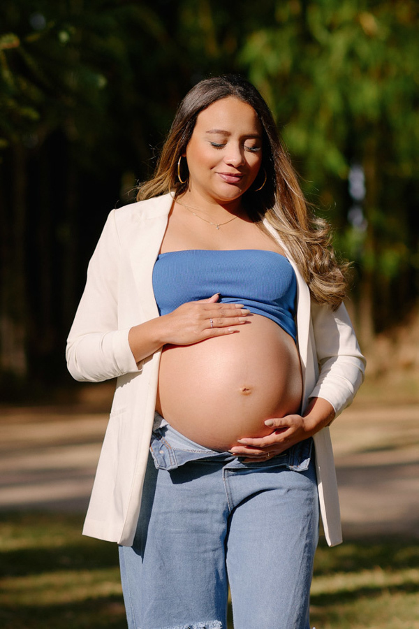 Fotografia de gestante em São José dos Campos. Lorrana sorri suavemente enquanto acaricia a barriga à espera de Pedro. O registro destaca a luz natural do Parque da Cidade, realçando a serenidade e a beleza da maternidade em um ensaio ao ar livre.