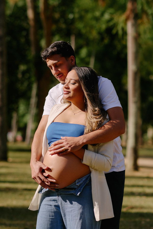 Ensaio gestante em São José dos Campos. Felipe abraça Lorrana por trás enquanto ambos repousam as mãos sobre a barriga à espera de Pedro. O casal posa de olhos fechados sob a luz do sol no Parque da Cidade, em um registro de profunda conexão e paz.