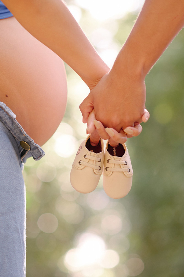 Detalhe do ensaio gestante em São José dos Campos. As mãos de Lorrana e Felipe dadas, segurando sapatinhos de bebê à frente da barriga. O close destaca a expectativa por Pedro em um cenário com luz suave e bokeh natural no Parque da Cidade.