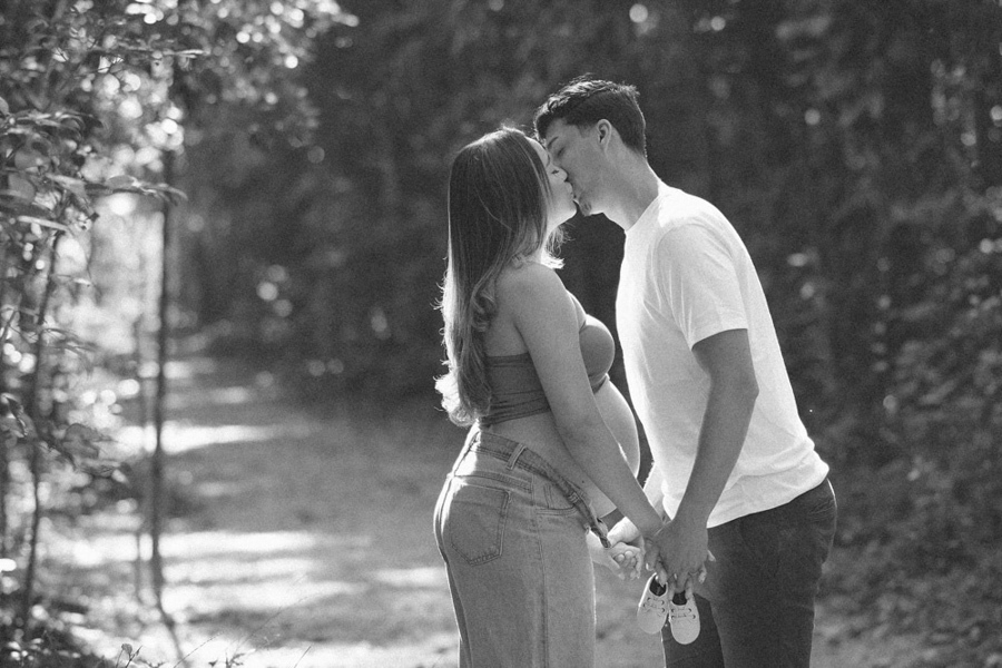 Foto de gestante em preto e branco em São José dos Campos. Lorrana e Felipe se beijam enquanto seguram os sapatinhos de Pedro no Parque da Cidade. O registro atemporal destaca a silhueta da barriga e a luz suave entre as árvores do ensaio.