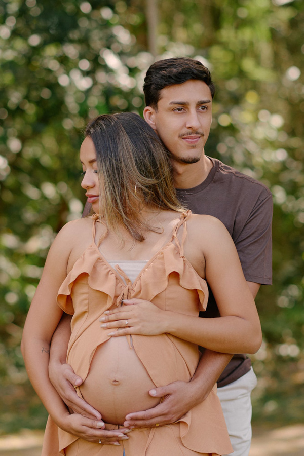 Ensaio gestante em São José dos Campos. Felipe abraça Lorrana por trás em um momento de ternura no Parque da Cidade. Ambos vestem tons terrosos, com as mãos suavemente posicionadas sobre a barriga à espera de Pedro, em meio à natureza de SJC.