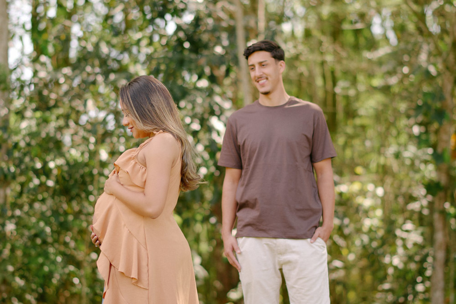 Fotografia de gestante em São José dos Campos. Lorrana em destaque acaricia a barriga, enquanto Felipe a observa sorridente ao fundo. O ensaio no Parque da Cidade utiliza o desfoque natural da vegetação para criar uma cena doce e espontânea em SJC.